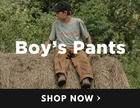 A young boy wearing carhartt pants sitting on a round hay bale